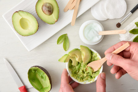 Hands preparing natural avocado skin care cream made in home kitchen with pieces of fruit on white...