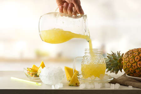 Person serving chilled pineapple juice from pitcher on kitchen bench with fruit and ice around. Frontview. Landscape composition.の写真素材