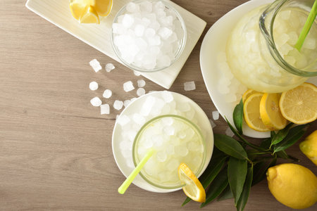 Background with lemon drink with a lot of ice in glass and pitcher on wooden table with fruit and a...