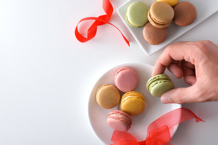 Hand tasting french macaroons from a white plate with different flavors on white table with red bows. top view.の写真素材