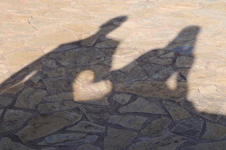Shadow on stone floor of a couple sitting on a wall holding hands creating a heart between their handsの写真素材