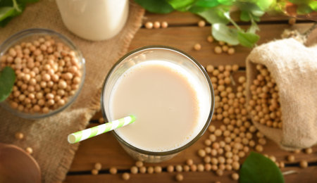 Detail of soy drink in glass cup with straw on wooden table with soybeans and leaves. top view.