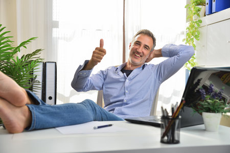 Happy smiling freelance worker in blue shirt making ok sign with hand and feet on the table looking straight working in white office with folders.の写真素材