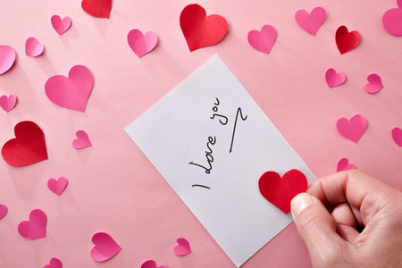 Romantic note with writing I love you on white paper on pink background with hand with a red heart cutout and many hearts around. Top view.の写真素材