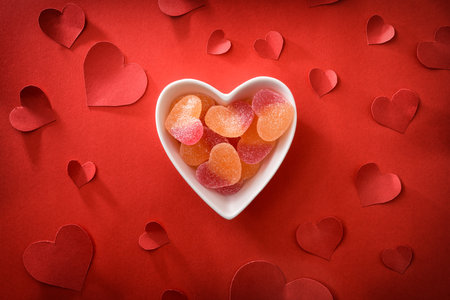 Conceptual love background with white ceramic container with heart-shaped candies on red background with red heart-shaped cutouts. Top view.の写真素材