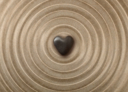 Black rounded heart-shaped stone on sand with concentric waves towards the center. Concept of mental and body health and well-beingの写真素材