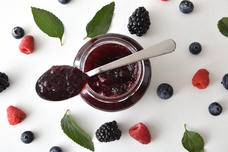 Background with spoon on jar with berry jam and blackberries, blueberries and raspberries on white table with leaves. Top view.の写真素材