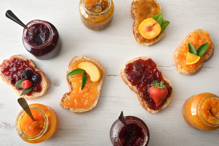slices of bread with jam of various flavors on a table with jars full of jam. Top view.