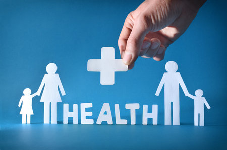 Family health background with white paper family cutout and hand holding health cross and letters forming the word health with blue isolated background. Front view.の写真素材