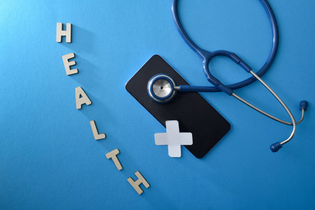 Concept of health and technological support with stethoscope on smartphone with medical cross and health message with wooden letters on blue isolated background. top viewの写真素材