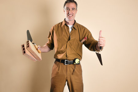 Smiling bricklayer making ok gesture with finger holding construction tools and materials and dressed in work clothes isolated on brown background. Front view.の写真素材