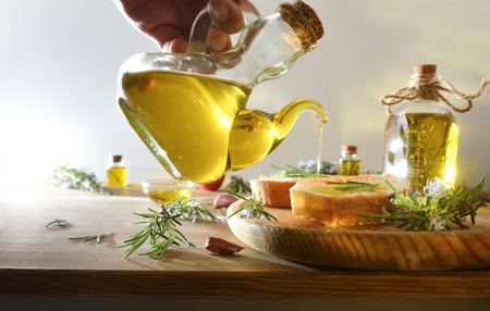 person dressing a slice of bread with a glass bottle of rosemary-essential oil on a wooden plate on a kitchen bench with rosemary sprigs and food on a white isolated background. Front view.の写真素材