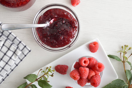 White wooden table with bowl full of raspberry jam and plate with berries and leaves. Top view.