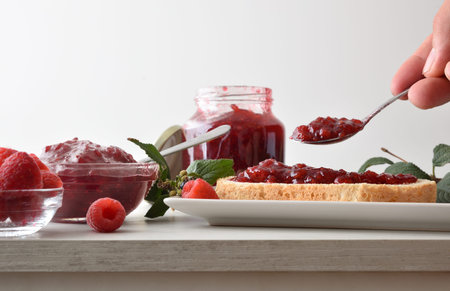 Person preparing toast with raspberry jam on white plate for breakfast at kitchen table with jars...