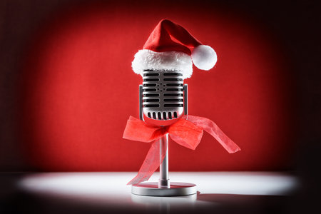 Retro microphone for performance with spotlight illuminating decorated with Christmas hat and red bow on white table isolated on red background. Front view.の写真素材