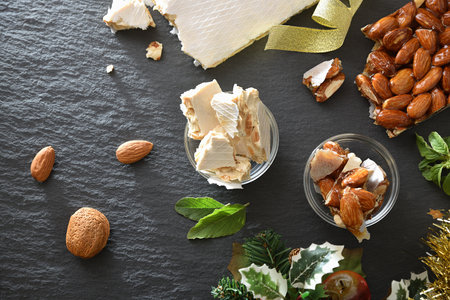 Two bars of hard nougat with almonds and pieces in bowls on a slate plate with natural almonds and Christmas decorations. Top view.の写真素材
