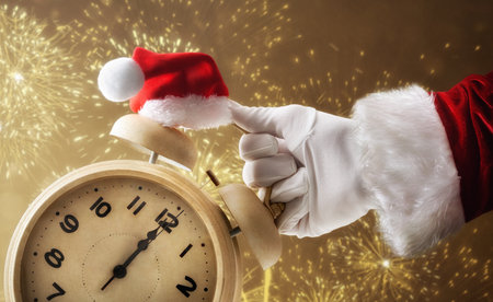 Santa's hand in a suit and glove holding a clock that shows 12 o'clock decorated with a Santa hat and golden fireworks in the background.の写真素材
