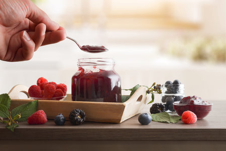 Hand filling spoon with forest fruit jam from a full jar on a wooden bench with berries and kitchen...