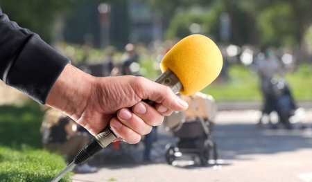 Hand in gray sweater holding metal handheld microphone with yellow foam windscreen to interview outside in a parkの写真素材
