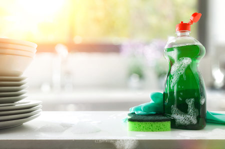 Dishwashing soap bottle, scouring pad and gloves on white bench with stack of dishes and kitchen...