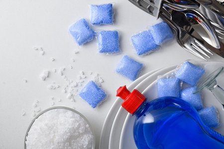 Detail of dishwasher products on white table like detergent in pods, salt in a bowl and rinse aid in a bottle with cutlery, plates, and glasses. Top view.の写真素材