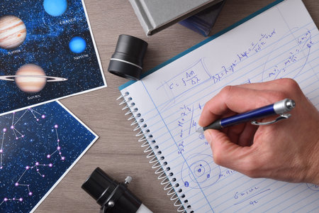 Student hand with pen making notes in a notebook on a wooden bench with photographs, books and parts of a telescope. Top view.の写真素材