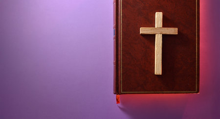 Detail of a brown leather Bible cover with a Christian cross on top and a red sparkle around it on a purple table. Top view.の写真素材