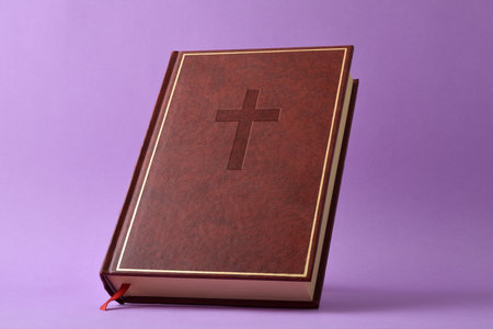 Detail of a closed Bible with a leather cover and a cross engraved with a red log tape isolated on a purple background.の写真素材