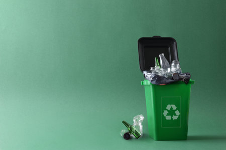 Green trash container with recycling symbol filled with glass objects and containers waste and other glass waste next to it on the ground isolated with green gradient background. Front view.の写真素材