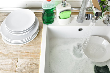 Detail of kitchen sink with white dishes and scouring pad with a running tap running water onto a wood effect tiled worktop with a stack of freshly washed dishes and cleaning products. Top view.の写真素材