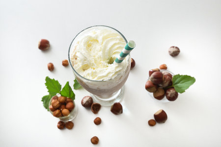 Glass with hazelnut smoothie with cream and green straw on white table with bowls full of hazelnuts and leaves around on isolated on white table. Top view.の写真素材