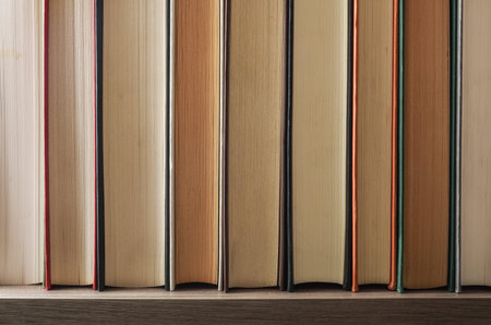 Detail of a row of books showing the front edge on a wooden shelf and dark isolated background....