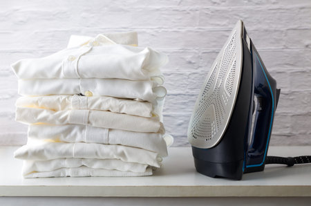 stack of white shirts on a white wooden cabinet with an iron next to it in an ironing room. Front view.の写真素材
