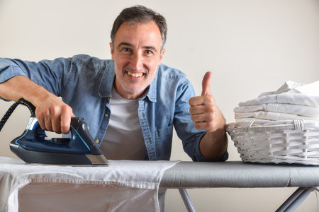 Detail of smiling elegant man ironing a white shirt on a gray ironing board with a basket full of freshly ironed shirts and ok gesture and white isolated background. Front view.の写真素材