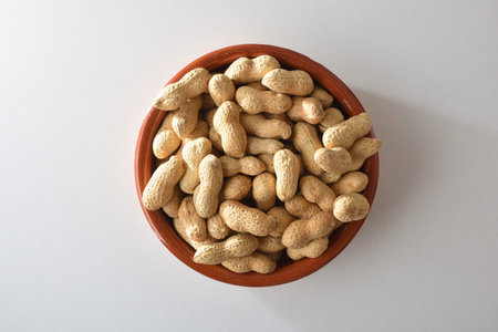 Heap of peanuts with skin in a brown clay bowl isolated on a white table. Top view.