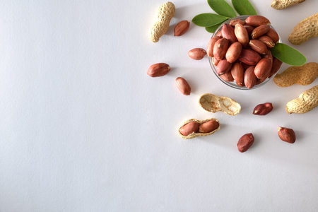 Heap of peeled peanuts with skin in a glass bowl isolated on a white table with leaves. Top view.
