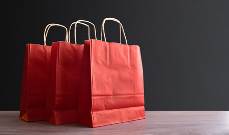 Pack of three empty red paper shopping bags with brown twisted paper handles on a wooden table and dark isolated background. Front view.の写真素材