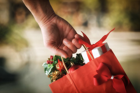 Christmas gift concept with hand holding red shopping bag with gifts and decorations in the outdoor commercial area. Front view.の写真素材