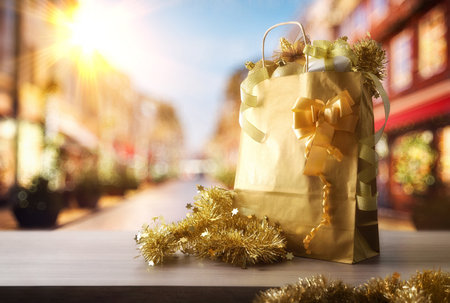 Christmas shopping concept with a golden gift bag containing gift packages and Christmas decorations on a wooden table in the middle of a street lined with shops on a sunny day.の写真素材