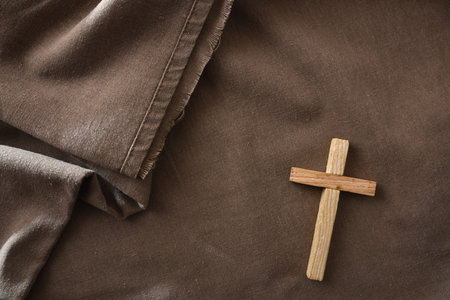Small wooden cross resting on brown fabric with soft folds. Warm, rustic composition representing...