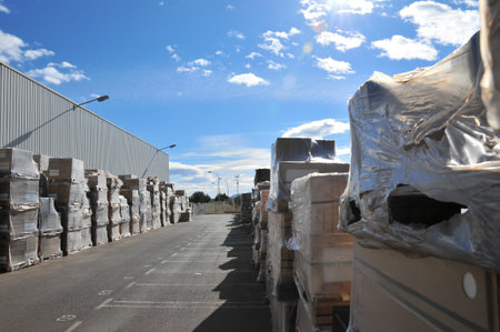 Outdoor industrial yard with pallets stacked with boxed ceramic tiles wrapped in plastic next to a...