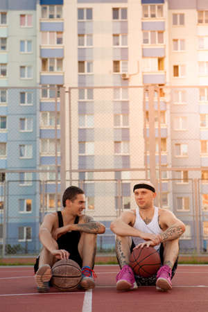 Two guys are sitting on the basketball court with balls, chatting and smiling.の写真素材