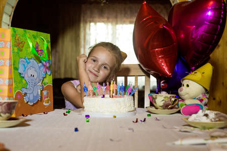 Little girl's birthday sitting at a table in front of the cake and looks at the camera.の写真素材