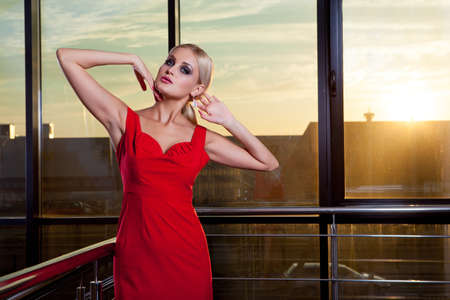 Young beautiful glamorous blonde girl in a red dress posing on the background of glass showcase. She looks away.の写真素材