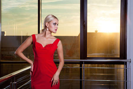 Young beautiful glamorous blonde girl in a red dress posing on the background of glass showcase. She looks away.の写真素材