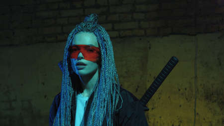 girl with dreadlocks and katanas in red glasses posing against a neon brick wallの写真素材