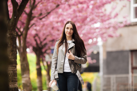 Girl in stylish outfit smiling and holding coffee on sakura background. Portrait of woman in jacket...