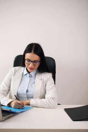 Cheerful brunette lady with important document friendly smiling and looking at camera while browsing laptop at desk in home officeの写真素材