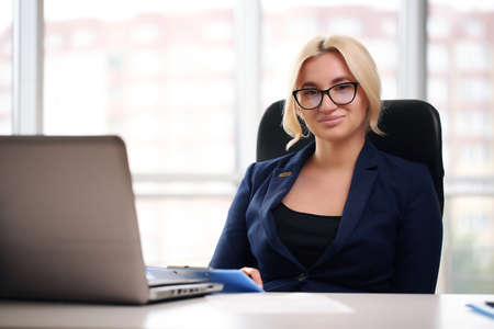 smiling blonde businesswoman with glasses using laptop in the officeの写真素材