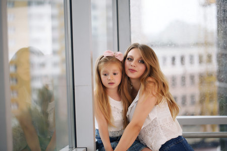 Mother and her baby girl playing hugging against the backdrop of a panoramic window